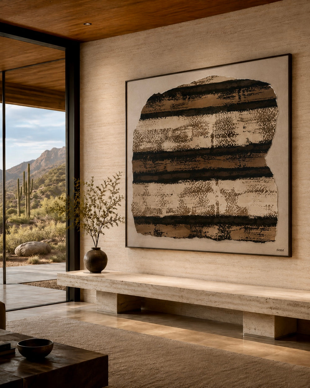 A large framed abstract wall art piece with horizontal textured bands in black, brown, and beige, displayed above a stone bench in a modern interior with a glass window overlooking a desert landscape.