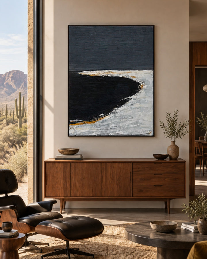 A framed abstract coastal painting hangs on a beige wall above a mid-century modern wooden credenza in a styled living room. The artwork features a dark navy and black curved landform with white and gold highlights.