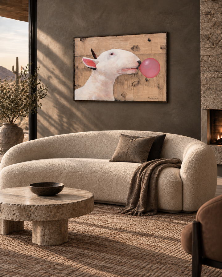 A framed wall art print of a white bull with a pink bubblegum bubble hanging above a curved cream boucle sofa in a modern living room with a stone coffee table and textured rug.