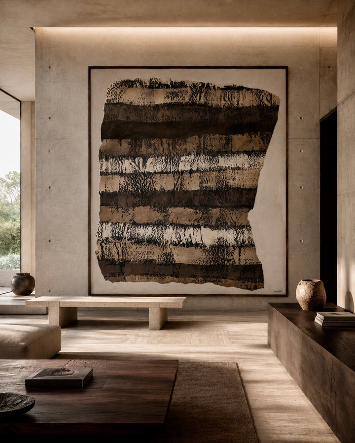 A large framed abstract wall art piece with horizontal textured brushstrokes in black, brown, beige, and white, displayed in a modern minimalist concrete living room.