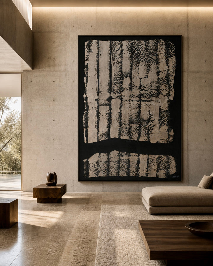 A large framed abstract black and off-white artwork hangs on a concrete wall in a modern minimalist living room with a beige sofa, wooden tables, and warm ambient lighting.